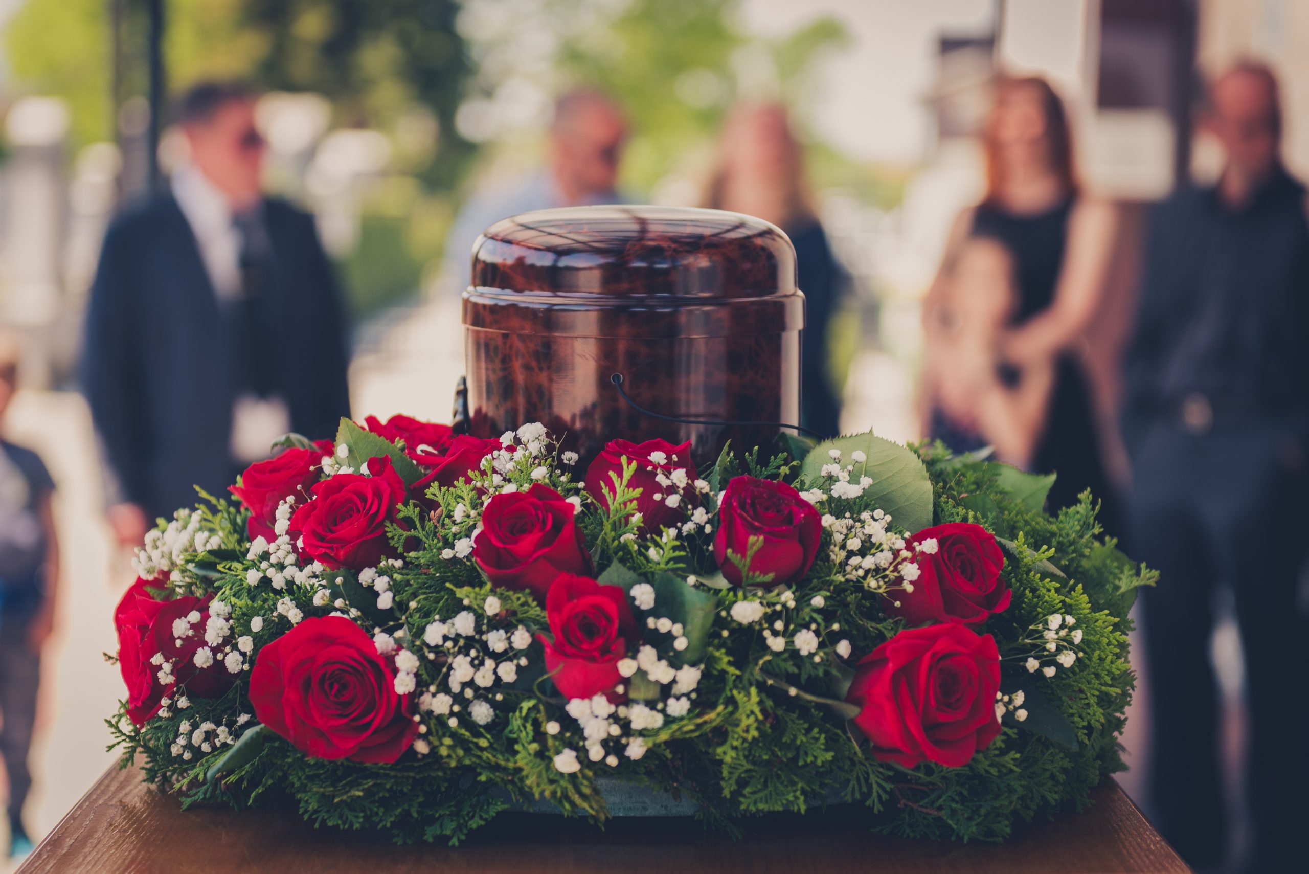 People attend a funeral with an urn in the foreground surrounded by a rose wreath People attend a funeral with an urn in the foreground surrounded by a rose wreath